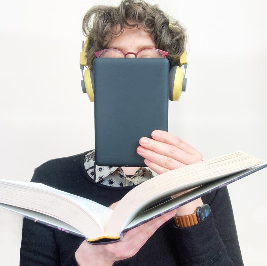 En kvinde med høretelefoner på holder en almindelig bog i den ene hånd og en e-bogslæser i den anden